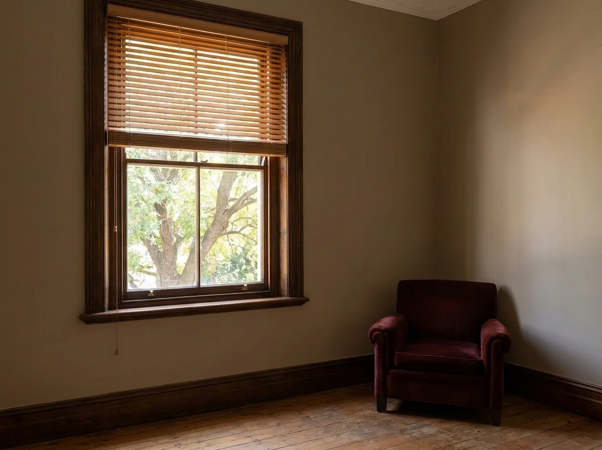 A tall heritage timber-framed window in an old Johannesburg house fitted inside the reveal with a timber venetian blind, its flat wooden slats tilted half open to angle dappled light from a large tree outside across a wooden floor